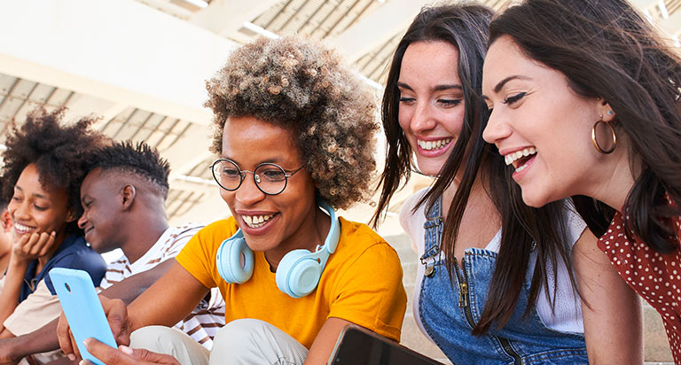 A group of young adults sit together on steps and are looking at something on their phones.