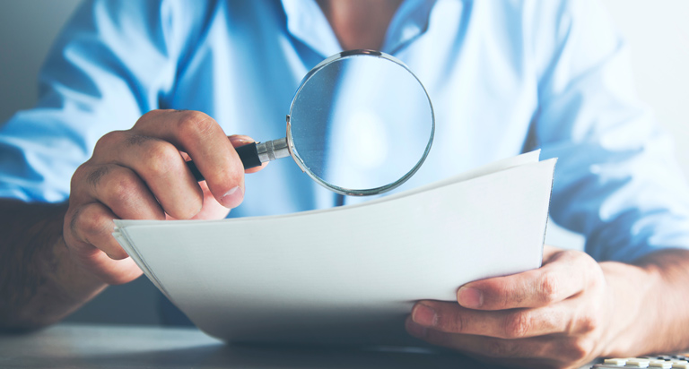 A man sits at a table with a calculator. He uses a magnifying glass in one hand to study pages of documents in the other hand.