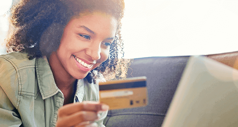 A smiling woman with dark hair lays on a couch while looking at a laptop and holding up a credit card in one hand.