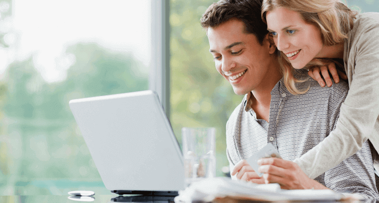 A man sits in front of his laptop smiling with a credit card in hand. A woman leans over his shoulder looking at the screen.